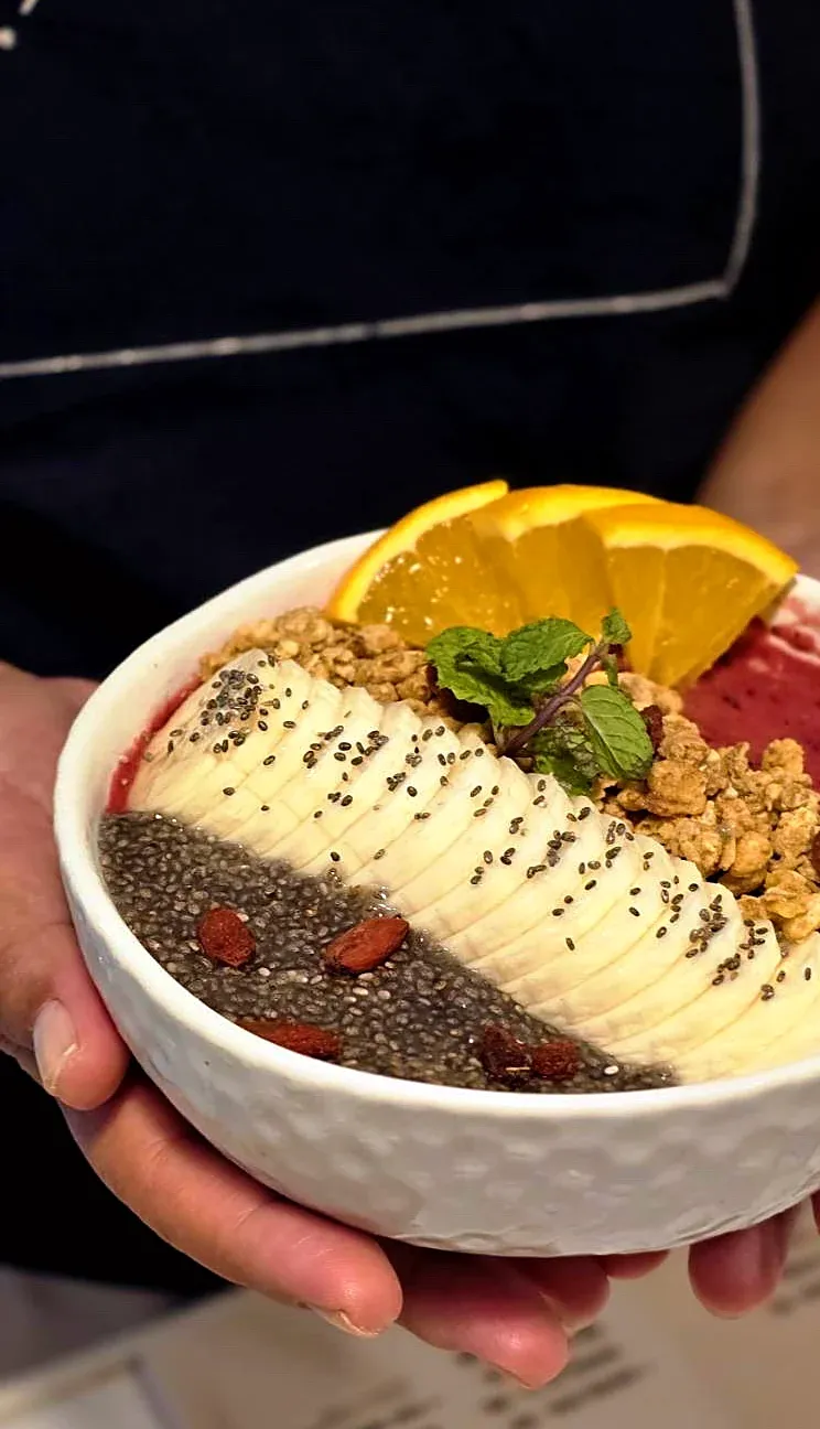 A Sway smoothie bowl held in both hands: acai and chia with sliced banana, granola, goji berries, mint and orange