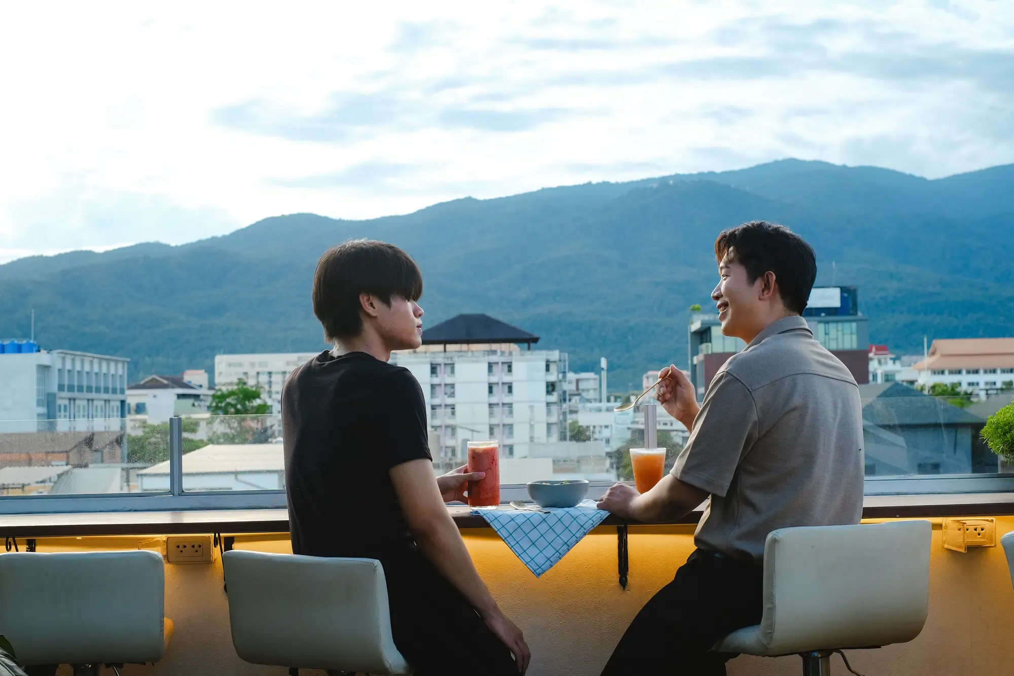 Two people at the rail on the Sway CNX rooftop in daylight, drinks on the counter, Chiang Mai and the mountains behind